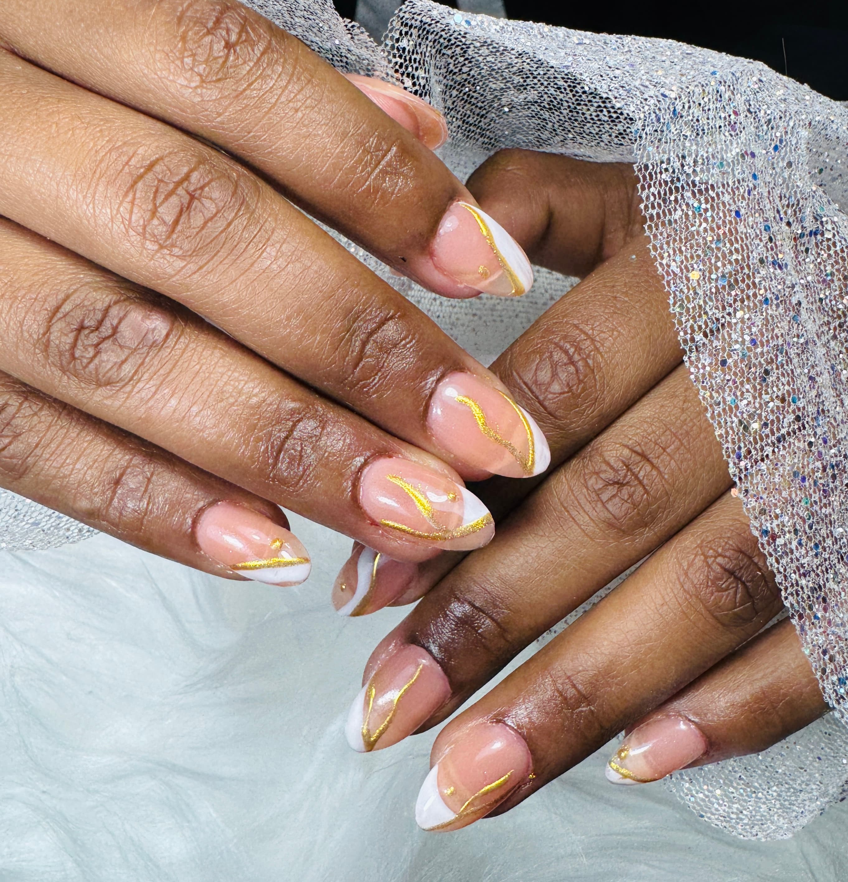 Manos con uñas almendradas nude, french blanco y líneas doradas, junto a un tejido plateado con brillo