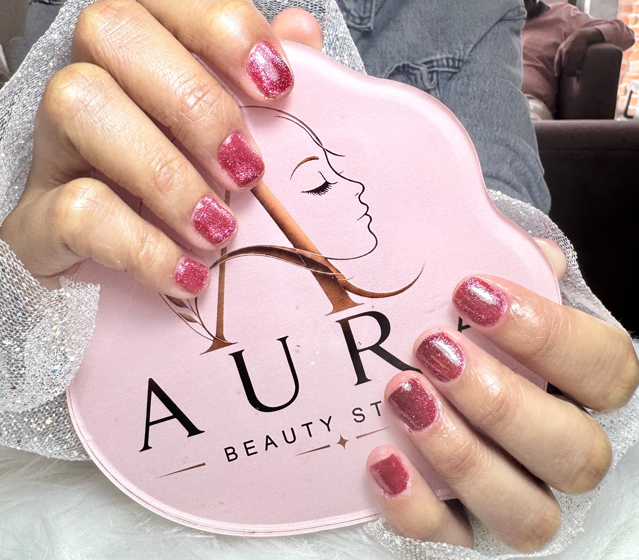 Manos con manicure corto en glitter rosa sosteniendo la paleta rosa con el logo de Aura Beauty Studio