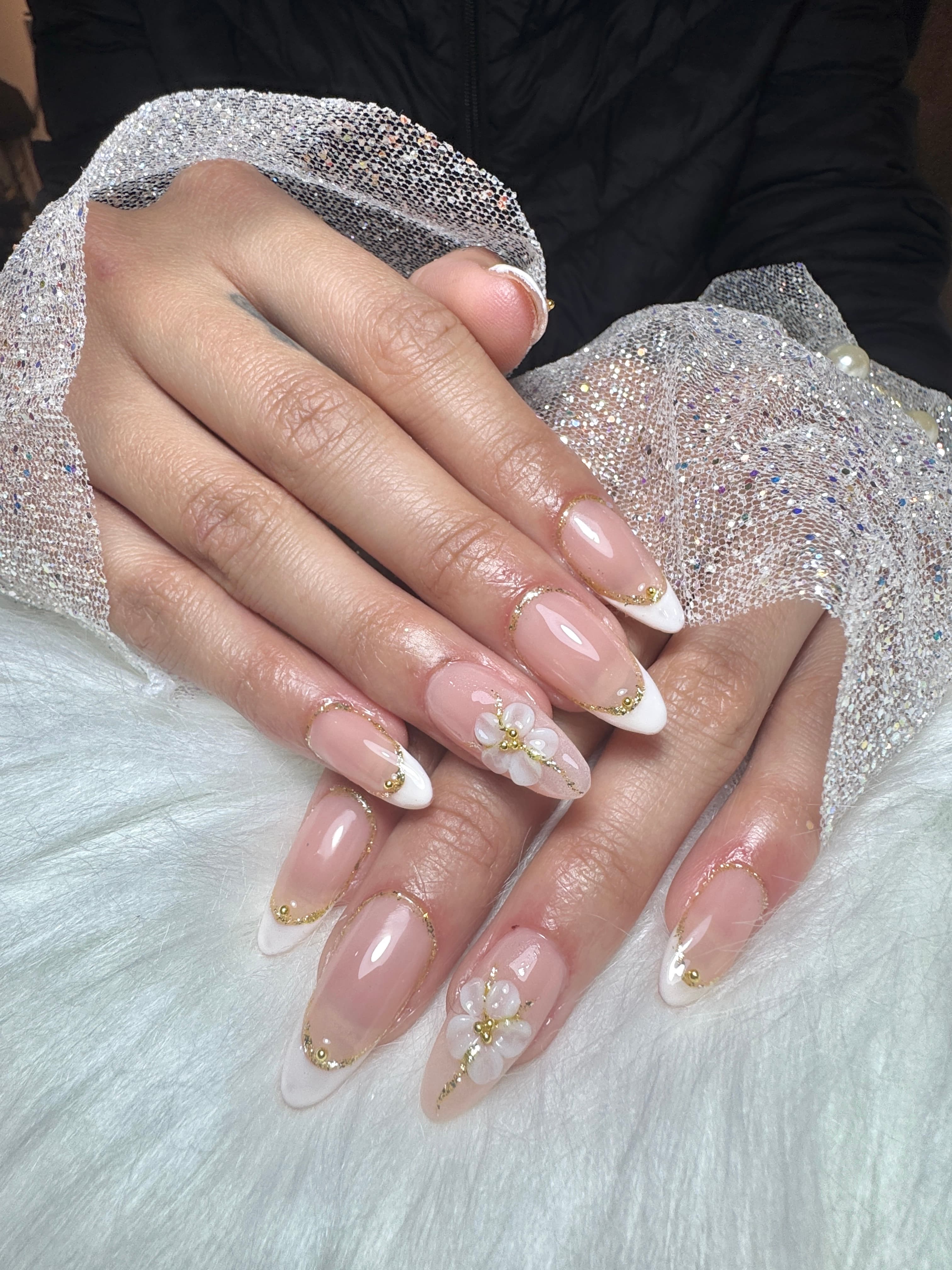 Uñas stiletto nude con french blanco, glitter dorado y flores 3D sobre un tejido plateado